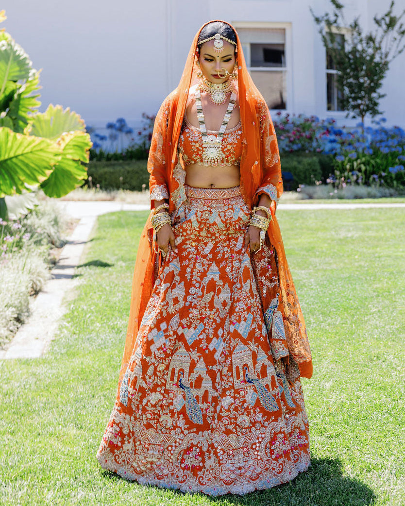 A woman stands outdoors on a green lawn in front of a white building, wearing an ornate orange and white embroidered traditional outfit with a matching sheer orange veil and heavy gold jewelry.
