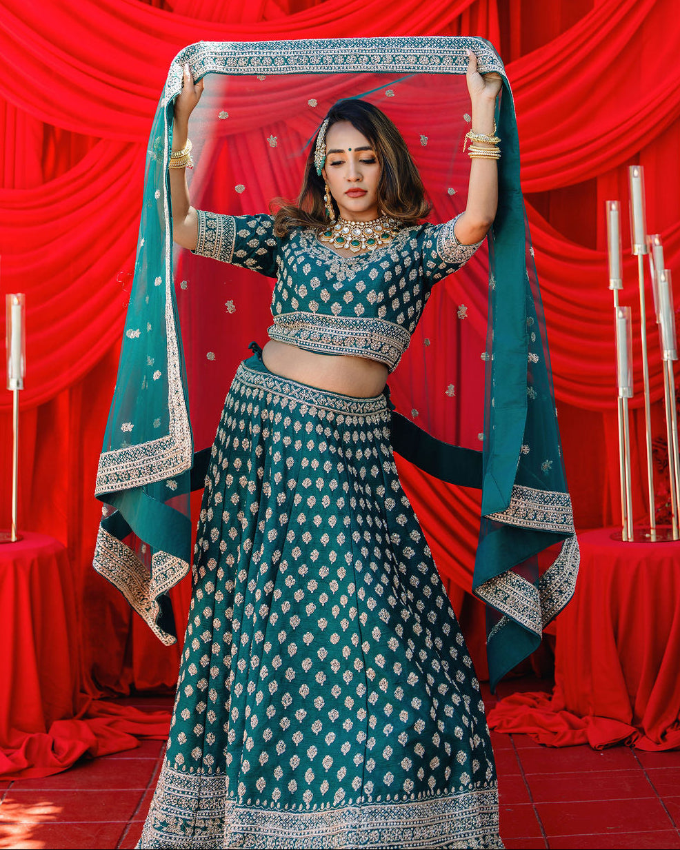 A woman modeling a teal blue and white printed lehenga choli set, holding up a matching dupatta against a red draped backdrop.