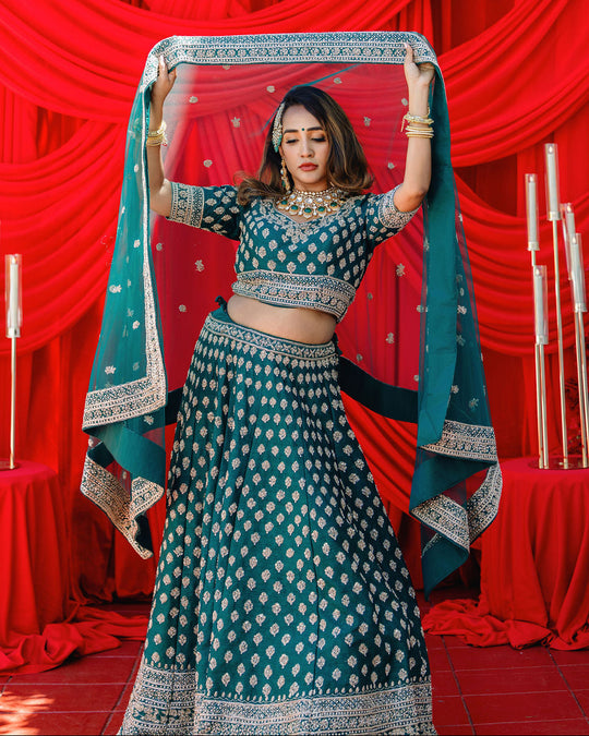 A woman modeling a teal blue and white printed lehenga choli set, holding up a matching dupatta against a red draped backdrop.
